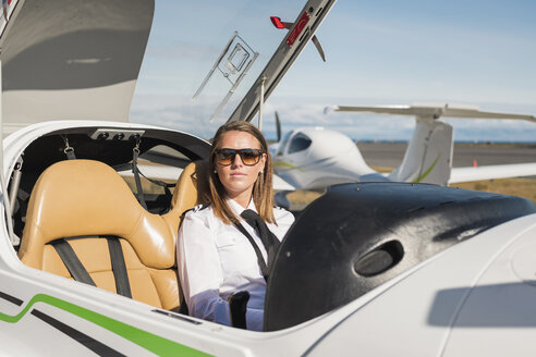 Porträt einer selbstbewussten Pilotin mit Sonnenbrille im Flugzeug sitzend gegen den blauen Himmel am Flughafen an einem sonnigen Tag - CAVF61809