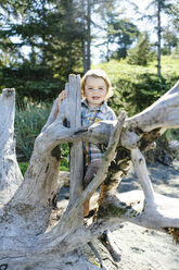 Portrait of cute smiling baby boy standing by driftwood at beach against trees during sunny day - CAVF61839