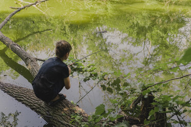 Junge in voller Länge beim Angeln, während er auf einem umgestürzten Baum in einem See im Wald kauert - CAVF61848