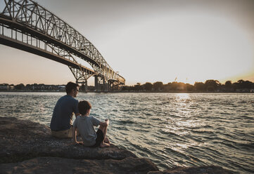 Father and son looking at view while sitting on rocks by sea against sky during sunset - CAVF61849