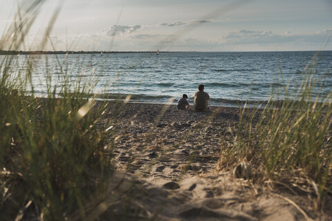 Rückansicht von Vater und Sohn am Strand sitzend bei Sonnenuntergang am Meer, lizenzfreies Stockfoto