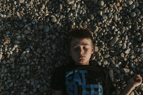 Draufsicht auf einen Jungen, der bei Sonnenuntergang mit geschlossenen Augen auf Kieselsteinen am Strand liegt, lizenzfreies Stockfoto