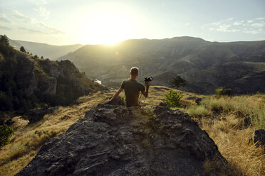 Rear view of man with camera looking at mountains while sitting on rock against sky during sunset - CAVF61890