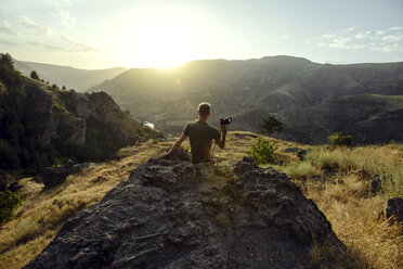 Rückansicht eines Mannes mit Kamera, der bei Sonnenuntergang auf einem Felsen sitzt und die Berge betrachtet - CAVF61890