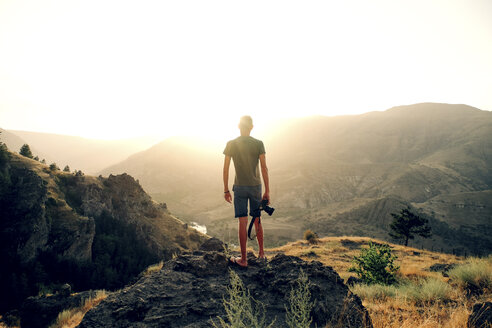 Rückansicht eines Mannes mit Kamera, der auf einem Felsen stehend bei Sonnenuntergang die Berge betrachtet - CAVF61892