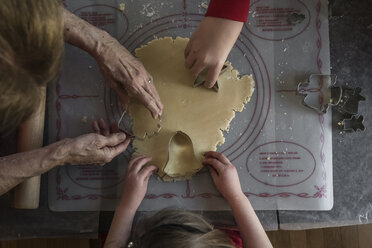 Ausgeschnittene Hände von Enkelkindern und Großmutter beim Schneiden von Teig mit Ausstechern auf dem Tisch zu Hause - CAVF61904