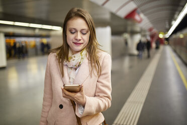 Österreich, Wien, Porträt einer lächelnden jungen Frau, die am Bahnsteig einer U-Bahn-Station auf ihr Smartphone schaut - ZEDF01947