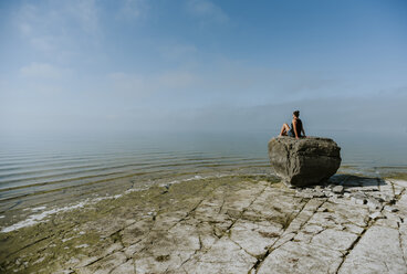 Frau schaut auf das Meer, während sie auf einem Felsen vor blauem Himmel sitzt - CAVF61989