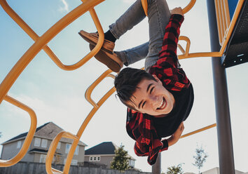 Porträt eines unbekümmerten, fröhlichen Jungen, der an einer Metalltreppe auf einem Spielplatz im Sommer hängt - CAVF61995
