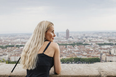 Rear view of woman with blond hair standing by retaining wall against cityscape - CAVF62001