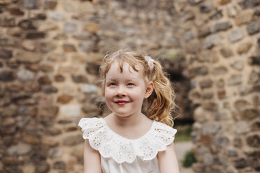 Cute smiling girl with blond hair looking away while sitting against old brick wall - CAVF62006
