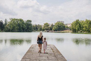 Rückansicht einer Mutter mit Tochter, die auf einem Steg über einem See gegen einen bewölkten Himmel in einer Stadt spazieren geht - CAVF62007