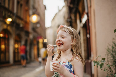 Glückliches Mädchen isst Essen, während sie auf der Straße vor Gebäuden in der Stadt steht - CAVF62015