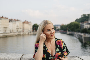 Frau hört Musik, während sie auf einer Fußgängerbrücke über den Fluss gegen den Himmel in der Stadt steht - CAVF62019