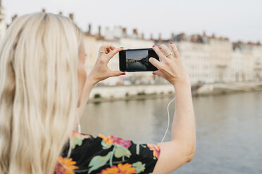 Seitenansicht einer Frau, die mit ihrem Mobiltelefon fotografiert, während sie auf einer Fußgängerbrücke über einen Fluss vor einem Gebäude in der Stadt steht - CAVF62021