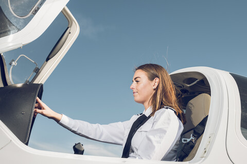 Seitenansicht einer Pilotin, die im Flugzeug sitzend das Steuerpult bedient, vor blauem Himmel am Flughafen, lizenzfreies Stockfoto