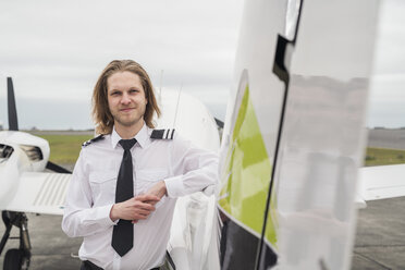 Porträt eines zuversichtlichen männlichen Piloten, der auf der Start- und Landebahn eines Flughafens bei bewölktem Himmel vor einem Flugzeug steht - CAVF62053