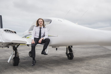 Porträt eines lächelnden männlichen Piloten, der in einem Flugzeug auf der Landebahn eines Flughafens vor einem bewölkten Himmel steht - CAVF62054