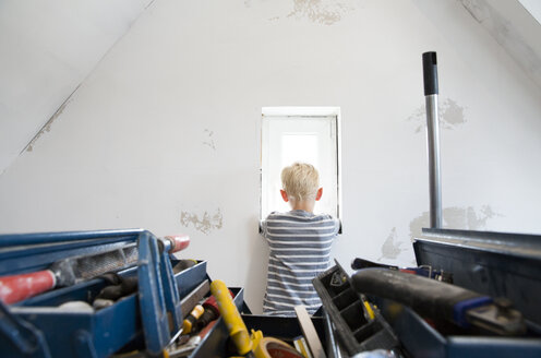 Junge schaut aus dem Fenster eines zu renovierenden Dachbodens mit Werkzeugkasten im Vordergrund - MFRF01186