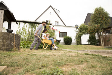 Playful man pushing wife and son sitting in wheelbarrow in garden - MFRF01277
