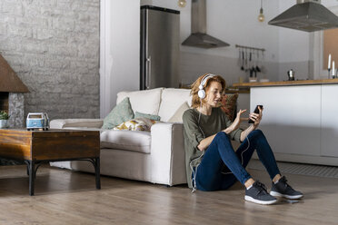 Young woman sitting on floor at home, using smartphone, wearing headphones - AFVF02532