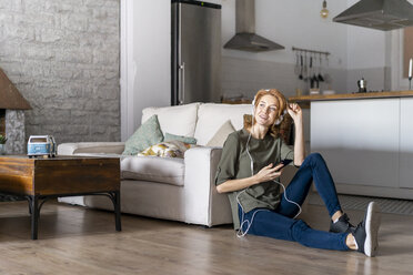 Junge Frau sitzt zu Hause auf dem Boden, benutzt ihr Smartphone und trägt Kopfhörer - AFVF02533