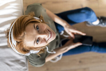 Junge Frau sitzt zu Hause auf dem Boden, benutzt ihr Smartphone und trägt Kopfhörer - AFVF02535