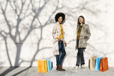 Portrait of two happy women with shopping bags standing at a wall - JRFF02763