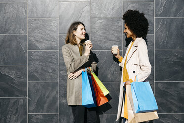Zwei glückliche Frauen mit Einkaufstaschen und Kaffee zum Mitnehmen stehen an einer Wand und unterhalten sich - JRFF02775