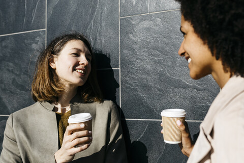 Zwei glückliche Frauen mit Kaffee zum Mitnehmen unterhalten sich an einer Wand, lizenzfreies Stockfoto