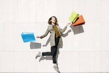Fröhliche Frau mit Einkaufstüten, die gegen eine Mauer springen - JRFF02783