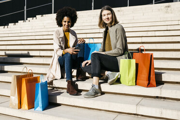 Portrait of two happy women with shopping bags sitting on stairs - JRFF02790