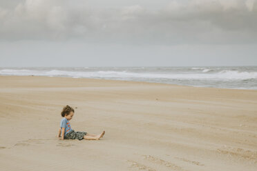 Side view of boy sitting on sand at beach against cloudy sky - CAVF62090