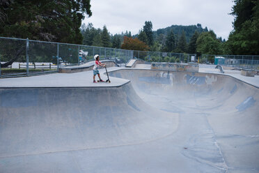 Side view of girl with push scooter standing on sports ramp at skateboard park - CAVF62117