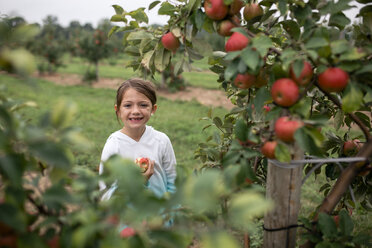 Porträt eines niedlichen lächelnden Mädchens, das bei Obstbäumen auf einem Bauernhof steht - CAVF62144