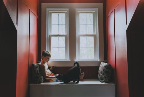 Seitenansicht des hemdlosen süßen Jungen, der mit einer Katze spielt, während er auf einem Alkoven-Fensterplatz zu Hause sitzt - CAVF62146