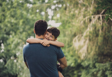 Rear view of father carrying happy cute son while standing against trees in forest - CAVF62150