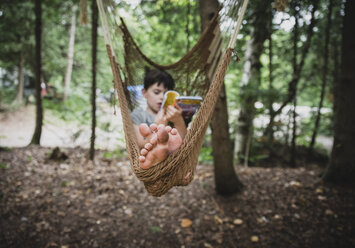 Full length of boy reading book while relaxing on hammock in forest - CAVF62152