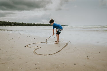 Junge macht Herzform auf Sand am Strand gegen bewölkten Himmel - CAVF62158