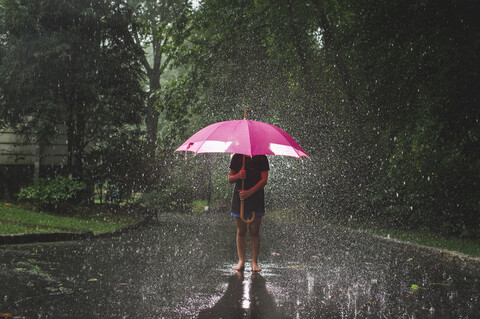 Mädchen mit Regenschirm, das bei Regen auf der Straße vor Bäumen steht, lizenzfreies Stockfoto