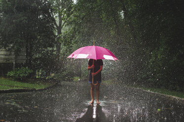 Mädchen mit Regenschirm, das bei Regen auf der Straße vor Bäumen steht - CAVF62163