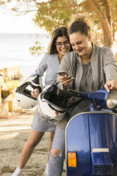 Smiling teenage girl standing by friend using phone while sitting on scooter - CAVF62174