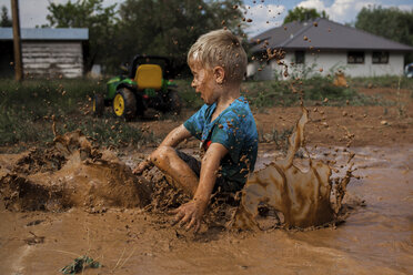 Side view of playful boy sitting on muddy puddle in yard - CAVF62206