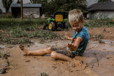 Side view of playful boy with arms outstretched sitting on muddy puddle in yard - CAVF62207