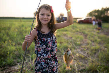 Porträt eines lächelnden Mädchens, das einen toten Fisch in einer Angelschnur auf einem Feld bei Sonnenuntergang hält - CAVF62218