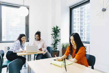 Businesswoman reading documents while colleagues discussing over laptop computer in office - CAVF62255