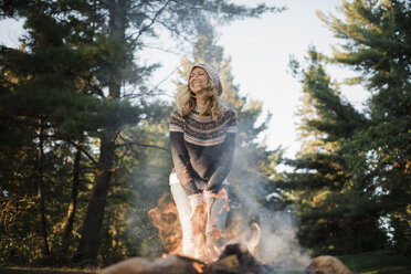 Glückliche Frau steht am Lagerfeuer vor Bäumen im Wald - CAVF62297