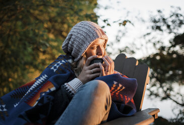 Thoughtful woman holding mug while sitting on chair in forest during sunset - CAVF62302