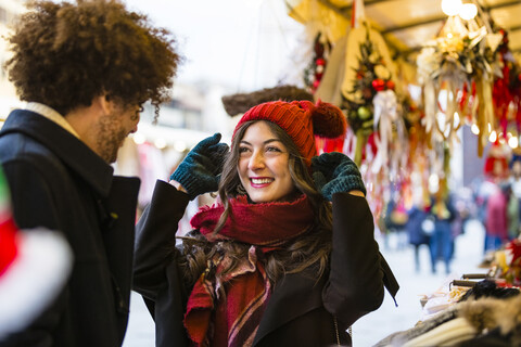 Glückliches junges Paar auf dem Weihnachtsmarkt mit Frau, die eine Wollmütze anprobiert, lizenzfreies Stockfoto