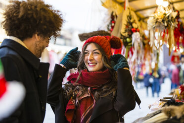 Glückliches junges Paar auf dem Weihnachtsmarkt mit Frau, die eine Wollmütze anprobiert - MGIF00301